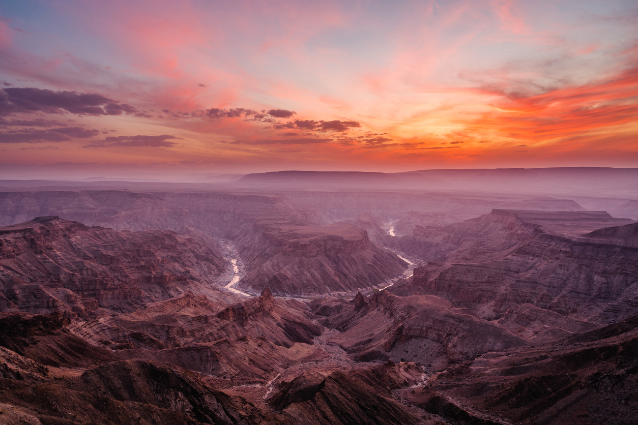 Wide view of Fish River Canyon at sunset with layered canyon walls, atmospheric haze, and the winding river below in Namibia.