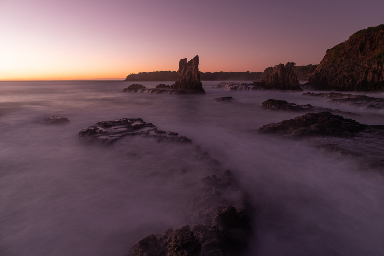 Cathedral Rocks rise from the ocean at dawn, surrounded by long-exposure waves beneath a soft pink and orange sky.