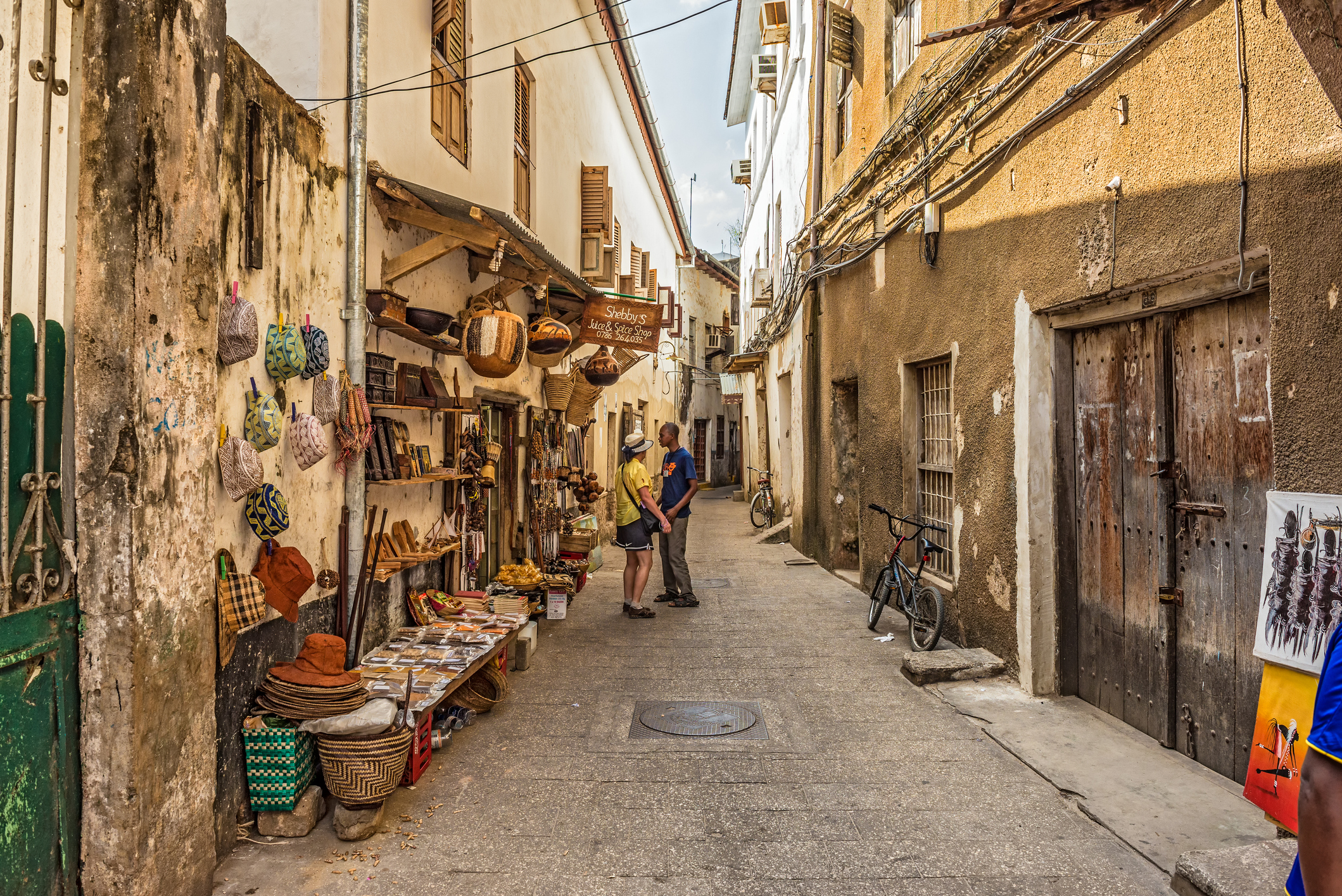 Narrow historic street in Stone Town, Zanzibar, with local craft shops, traditional baskets, and pedestrians exploring the old town.