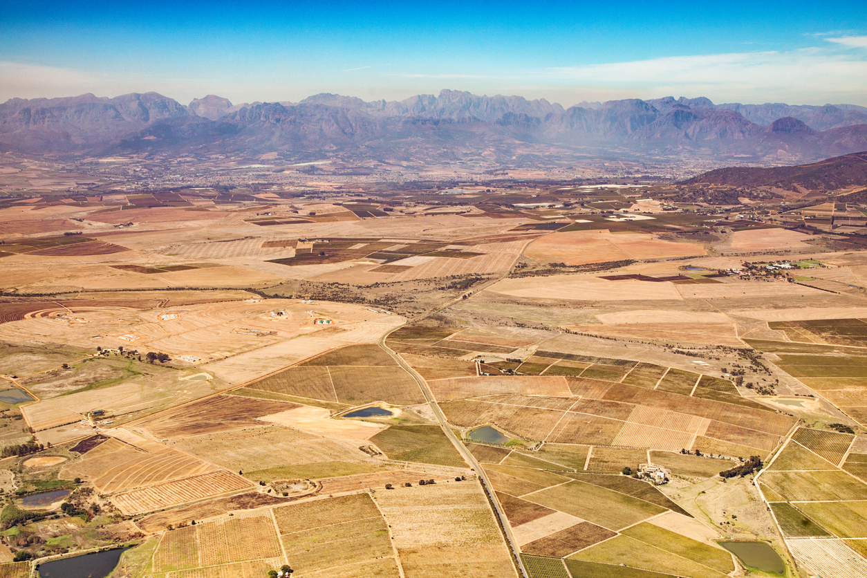 Aerial panorama of agricultural fields and vineyards near Wellington, South Africa, with geometric farm patterns, rural roads, irrigation dams, and distant Cape Fold mountains.