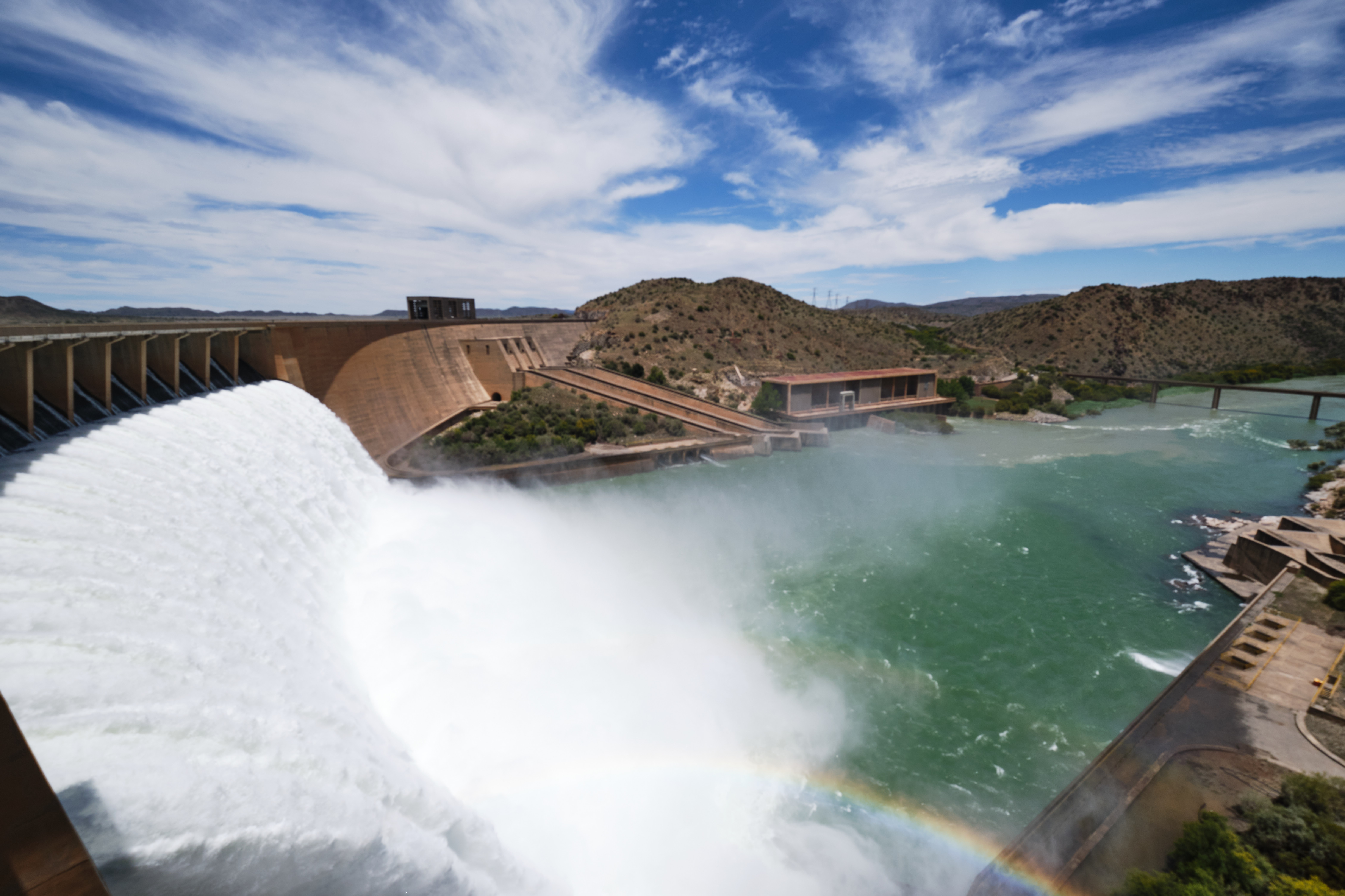 Water cascading over the Gariep Dam spillway into the Orange River below, with surrounding semi-arid hills and reservoir views in the Eastern Cape and Free State border region.