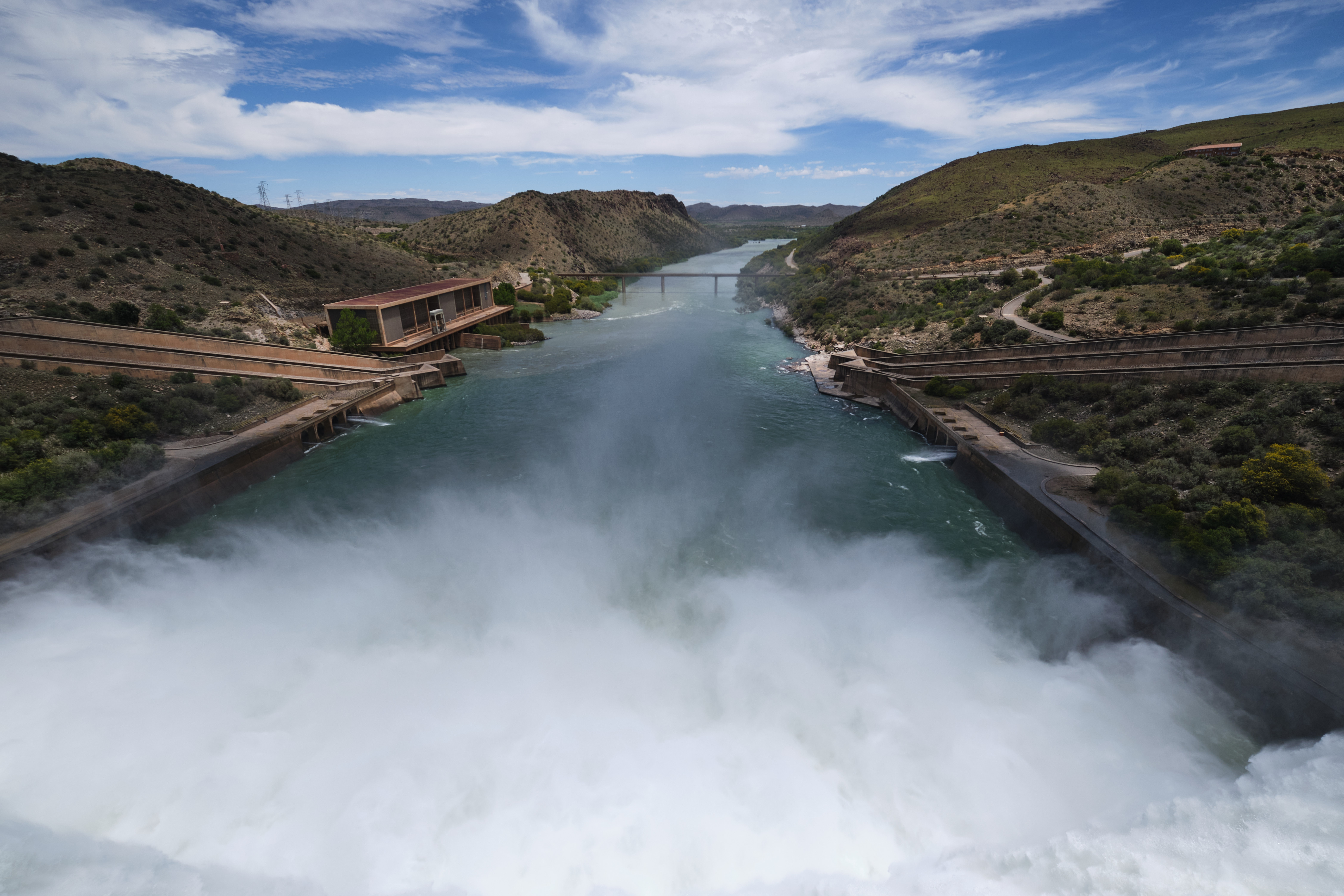 View downstream from Gariep Dam showing spillway spray, the Orange River channel and surrounding semi-arid hills below the dam wall.