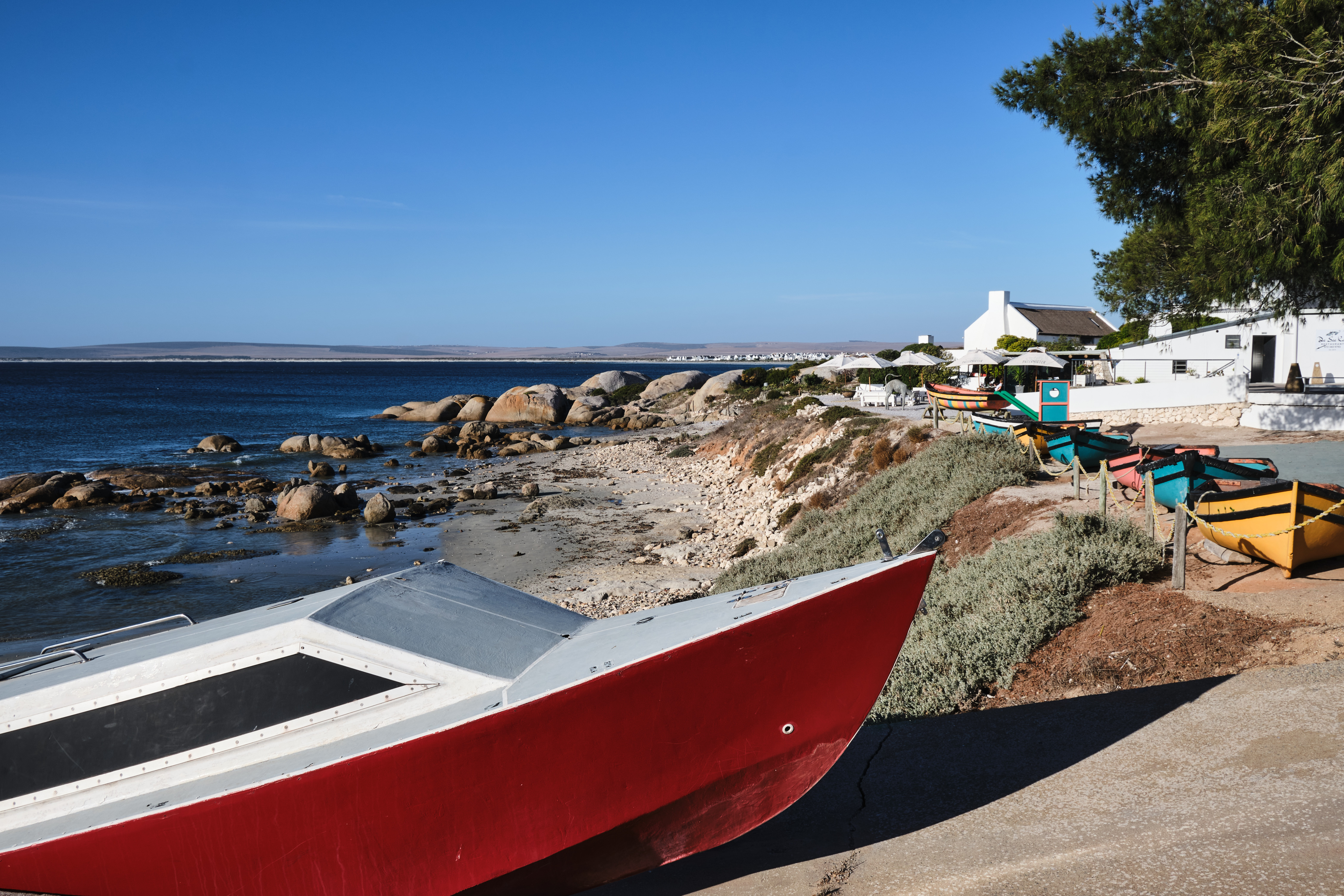 Red and brightly coloured fishing boats rest beside a rocky Atlantic shoreline with whitewashed buildings and coastal restaurants in Paternoster, Western Cape, South Africa.