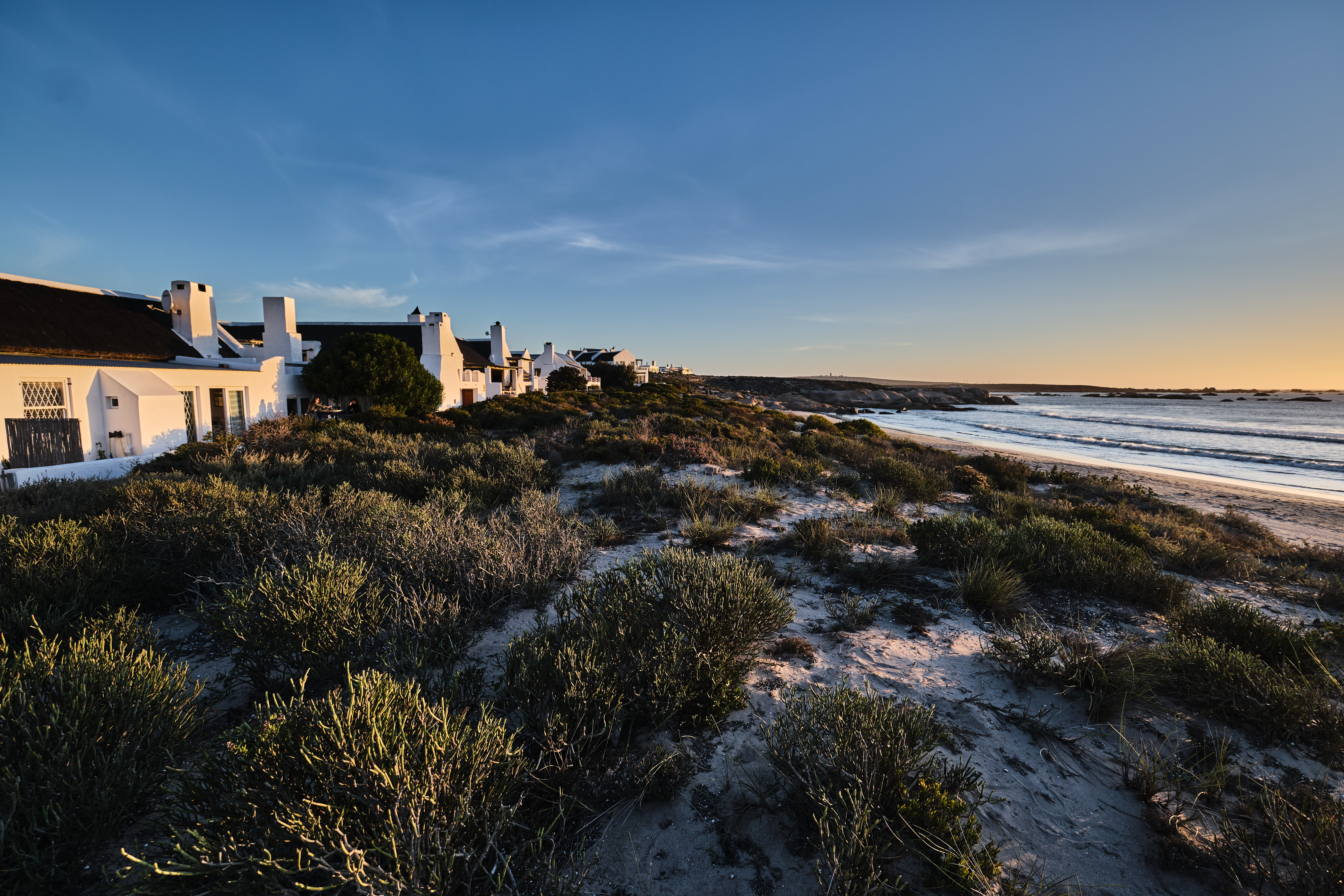 Whitewashed coastal cottages overlook a sandy beach and rocky shoreline at sunset in Paternoster, Western Cape, South Africa.