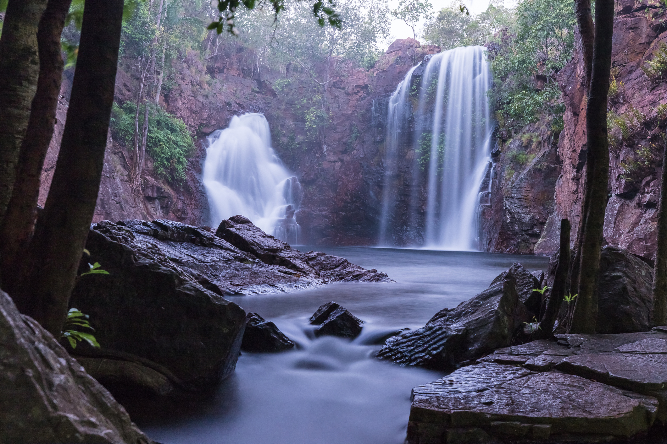 Atmospheric early morning view of the twin waterfalls at Florence Falls, framed by dark rocks and tropical trees, with silky flowing water entering the plunge pool in Litchfield National Park, Northern Territory, Australia.