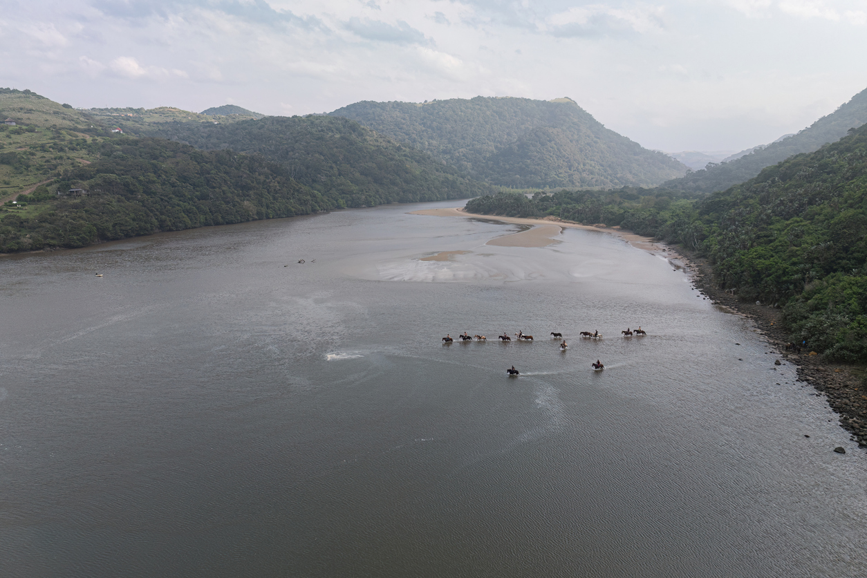 Aerial view of horse riders crossing the Mngazi River estuary surrounded by indigenous forest and sandbanks on South Africa's Wild Coast.