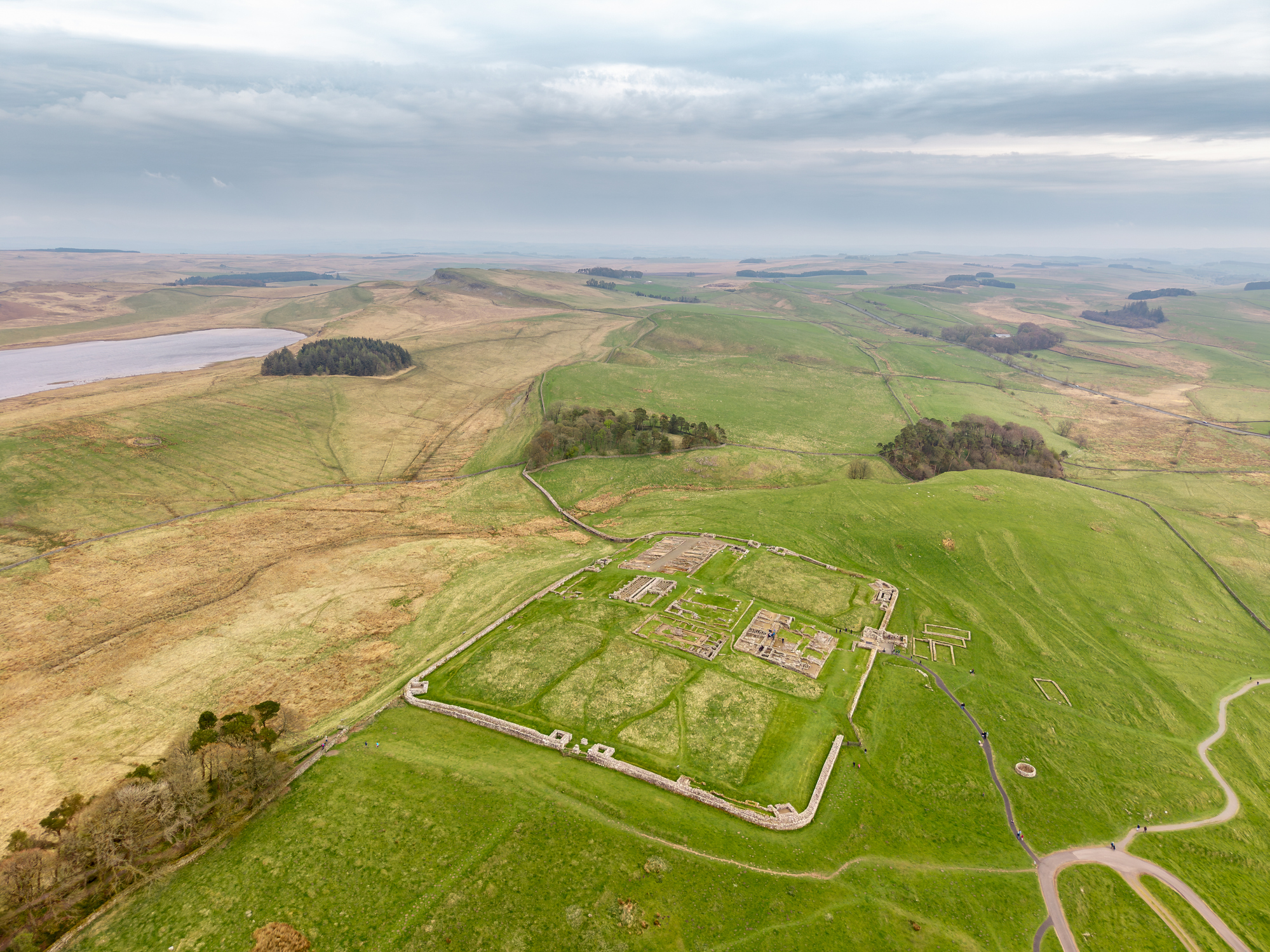 Drone view of the rectangular ruins and surviving stone foundations of Housesteads Roman Fort, surrounded by green hills and open Northumberland countryside.