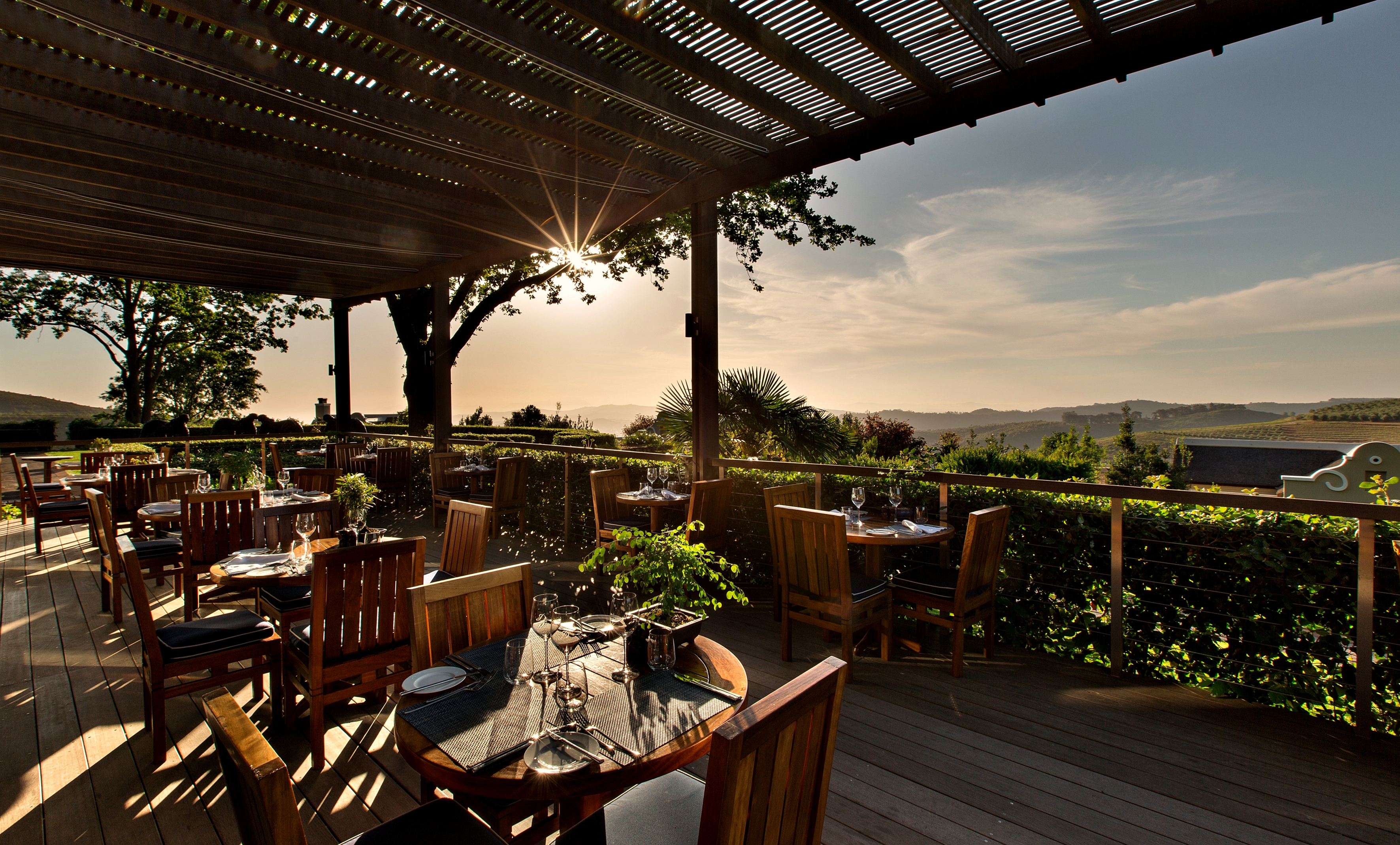 Outdoor terrace restaurant at Delaire Graff Estate overlooking vineyard-covered hills of the Cape Winelands at sunset.