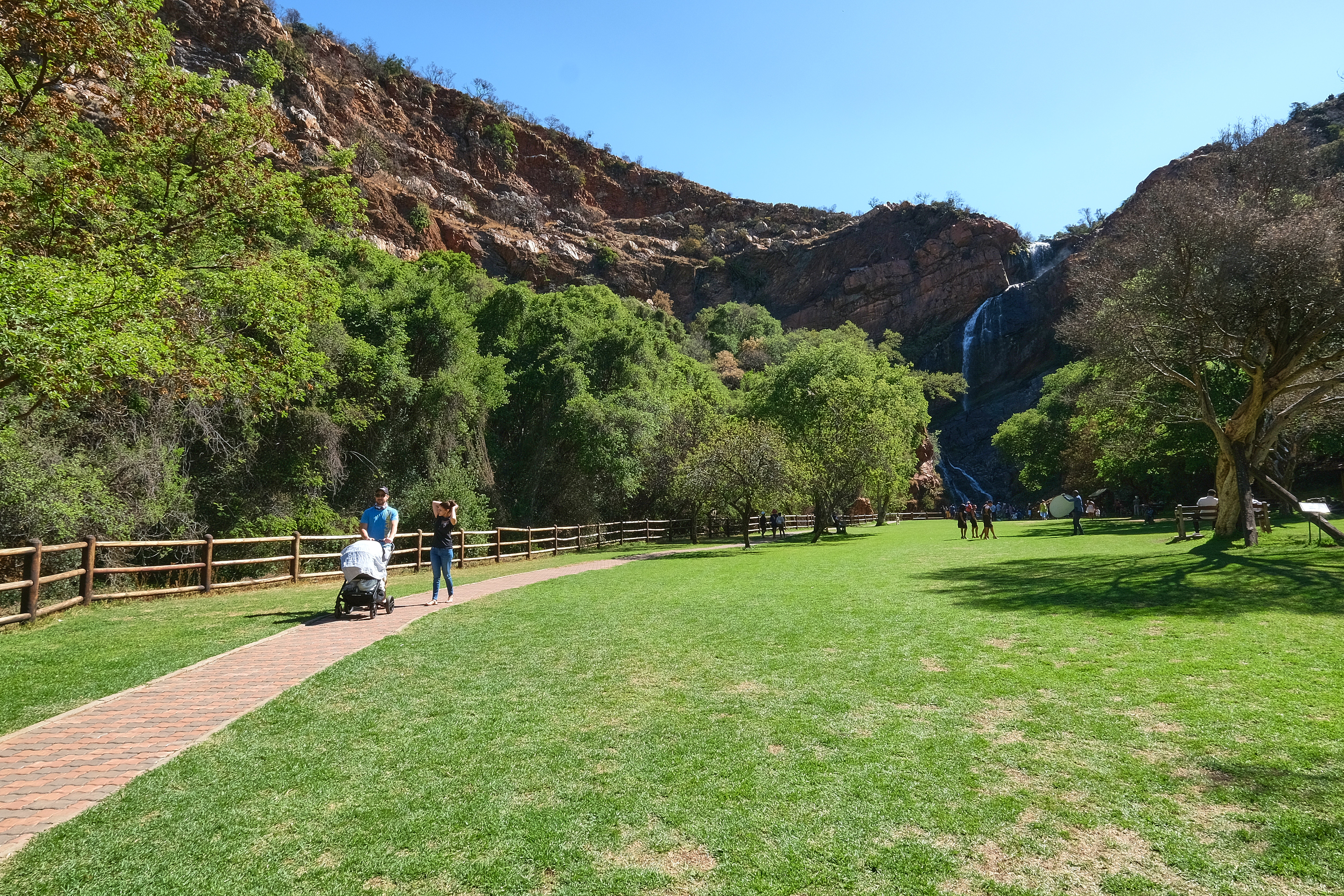 Visitors walking across open lawns beneath Witpoortjie Waterfall in Walter Sisulu National Botanical Garden, Roodepoort.