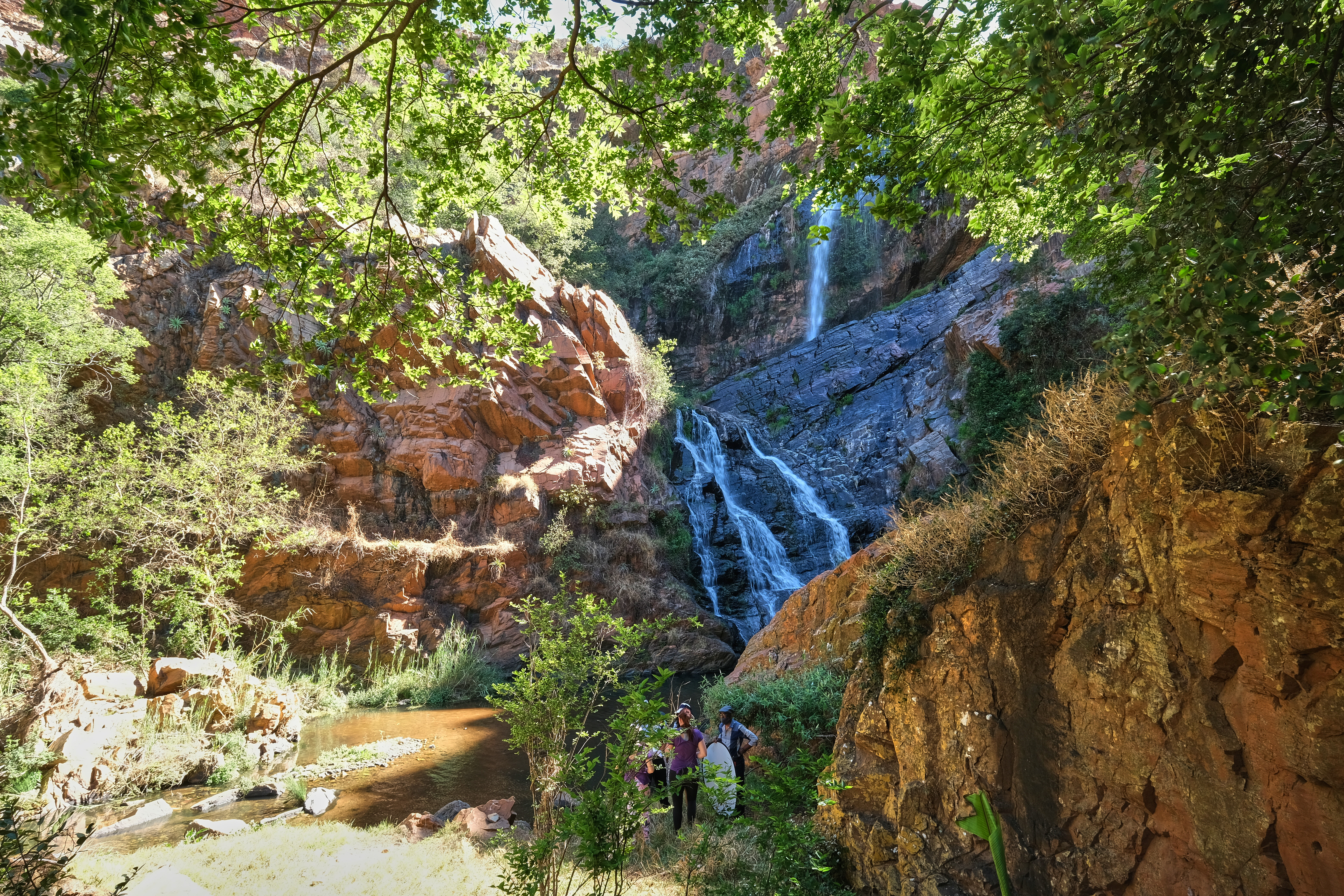 Visitors standing beside Witpoortjie Waterfall beneath red quartzite cliffs in Walter Sisulu National Botanical Garden, Roodepoort.