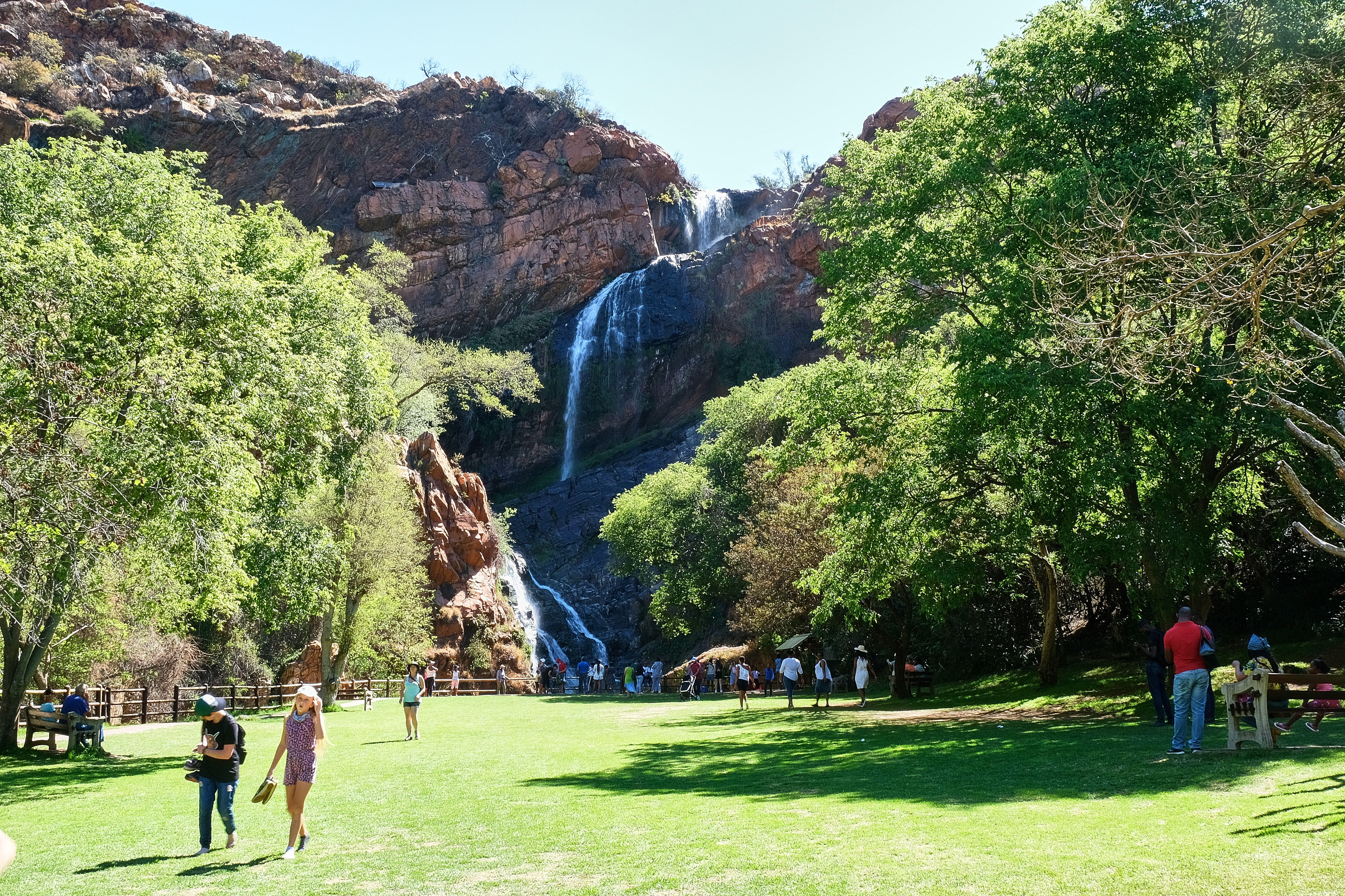 Visitors gather beneath the waterfall at Walter Sisulu National Botanical Garden in Roodepoort, surrounded by lawns, indigenous trees and rocky cliffs.