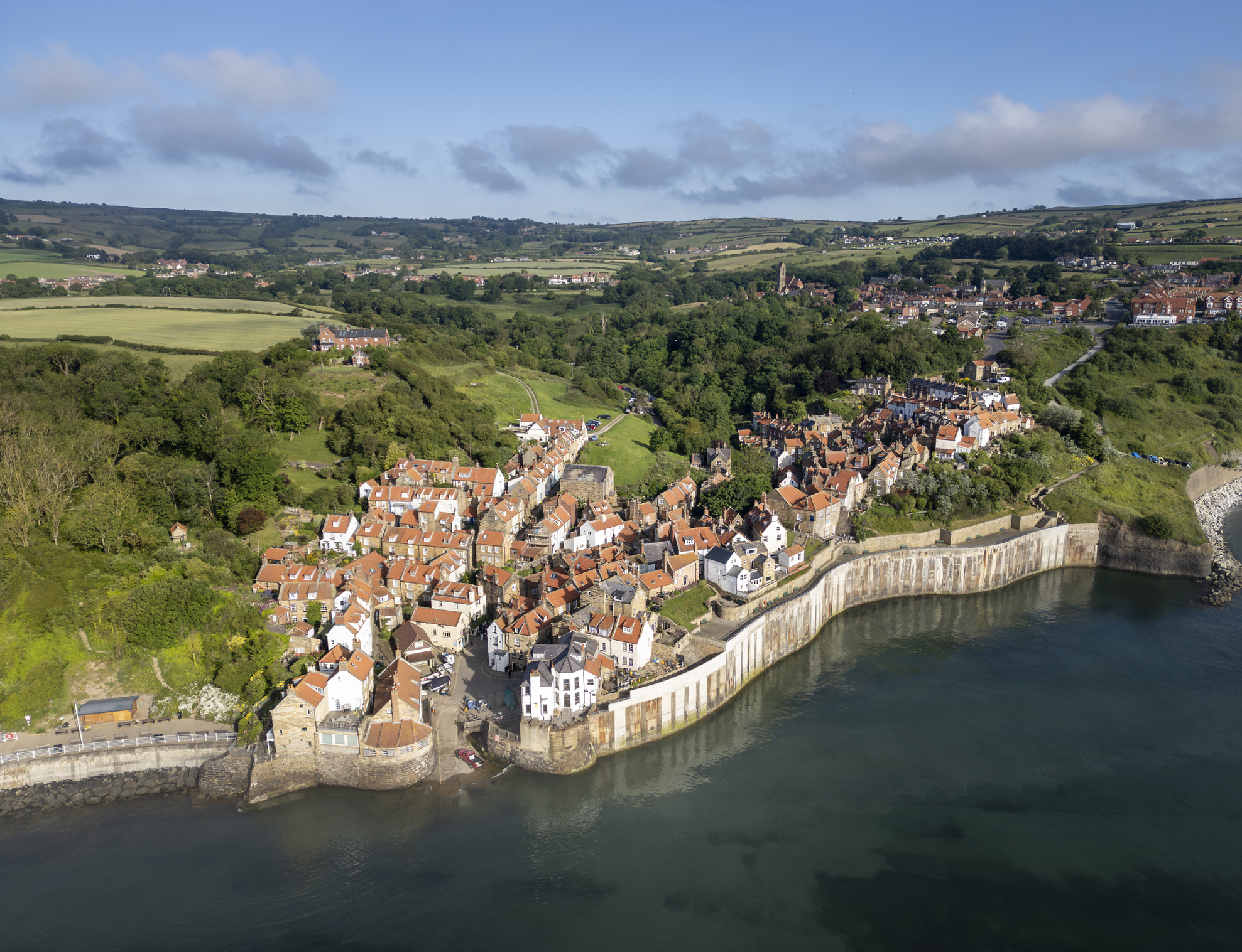 Aerial view of Robin Hood's Bay at high tide, showing red-roofed fishing cottages, the curved seawall and green hills along the North Yorkshire coast.