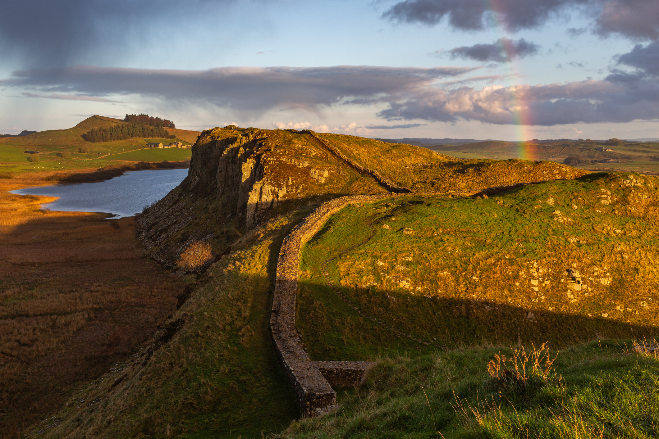 Hadrian's Wall curving along the rocky escarpment above Crag Lough in Northumberland, England, with golden evening light and a faint rainbow.