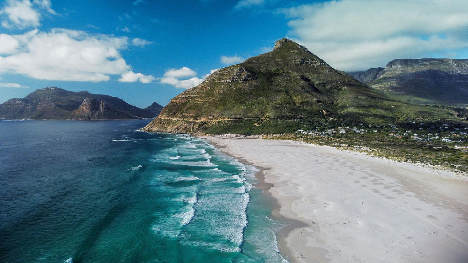 Aerial view of Noordhoek Beach with long white sand, turquoise waves, and Chapman’s Peak rising behind on the Cape Peninsula.