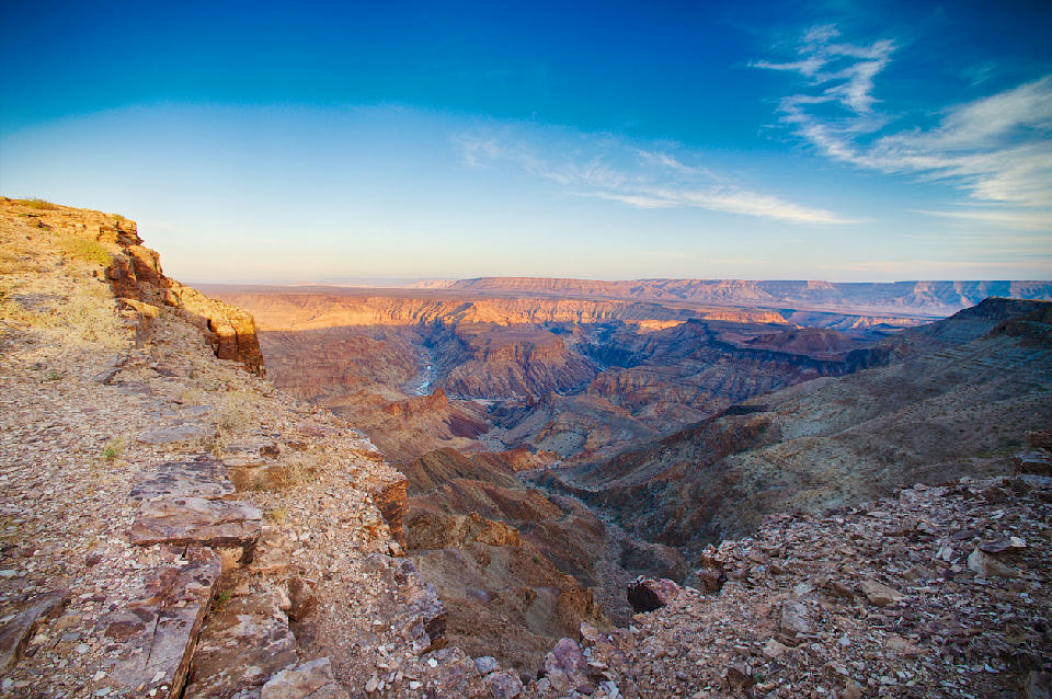 Cliff-edge sunrise view across Fish River Canyon in southern Namibia showing layered canyon walls, deep river valleys, and expansive desert escarpments beneath a blue sky.