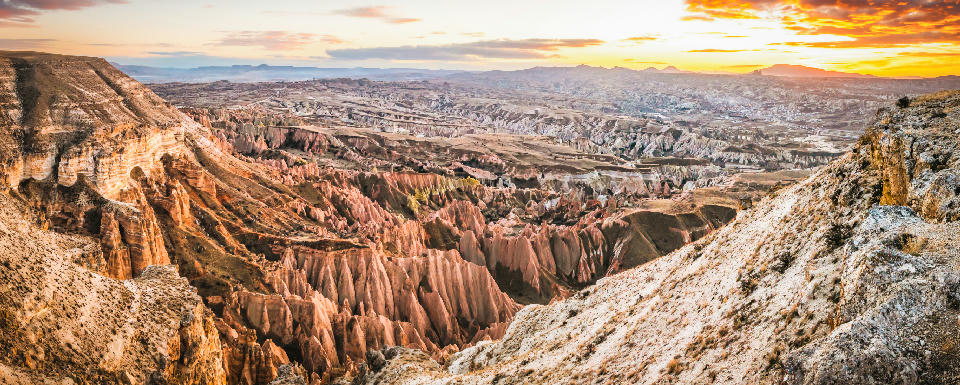 Panoramic sunset view over Red Valley in Cappadocia, Turkey, featuring colourful volcanic cliffs, deep canyons, fairy chimneys, and layered rock formations stretching across the landscape.