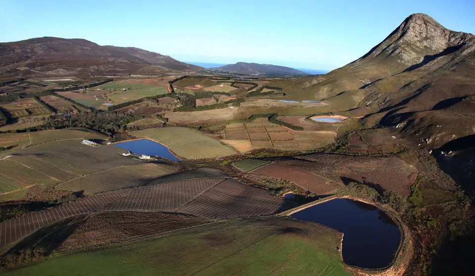 Aerial landscape including Creation Wines within the Hemel-en-Aarde Valley, showing vineyards, reservoirs, mountain ridges, and rolling terrain near Hermanus, South Africa.