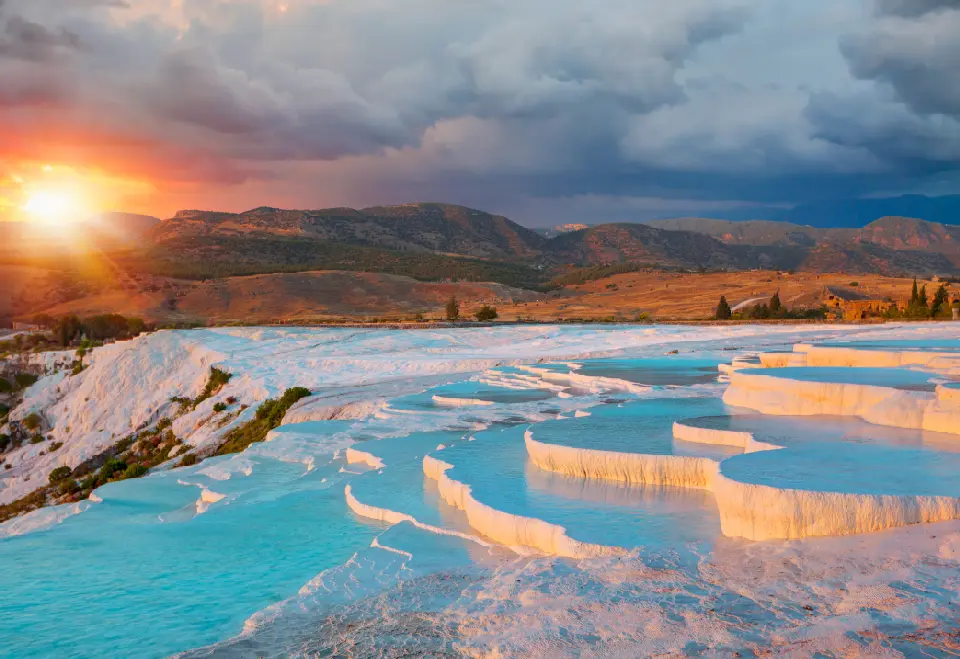 White travertine terraces filled with turquoise thermal water at Pamukkale, Turkey, illuminated by a dramatic sunset over the surrounding mountains.