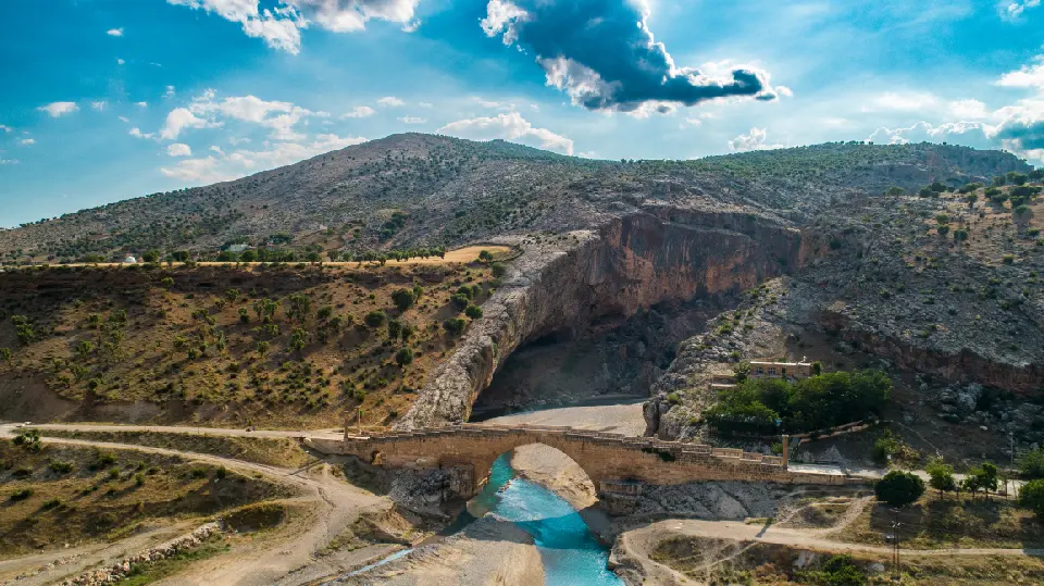 Aerial view of the ancient Roman Cendere Bridge crossing a turquoise river beneath steep rocky cliffs in Adıyaman, Turkey.