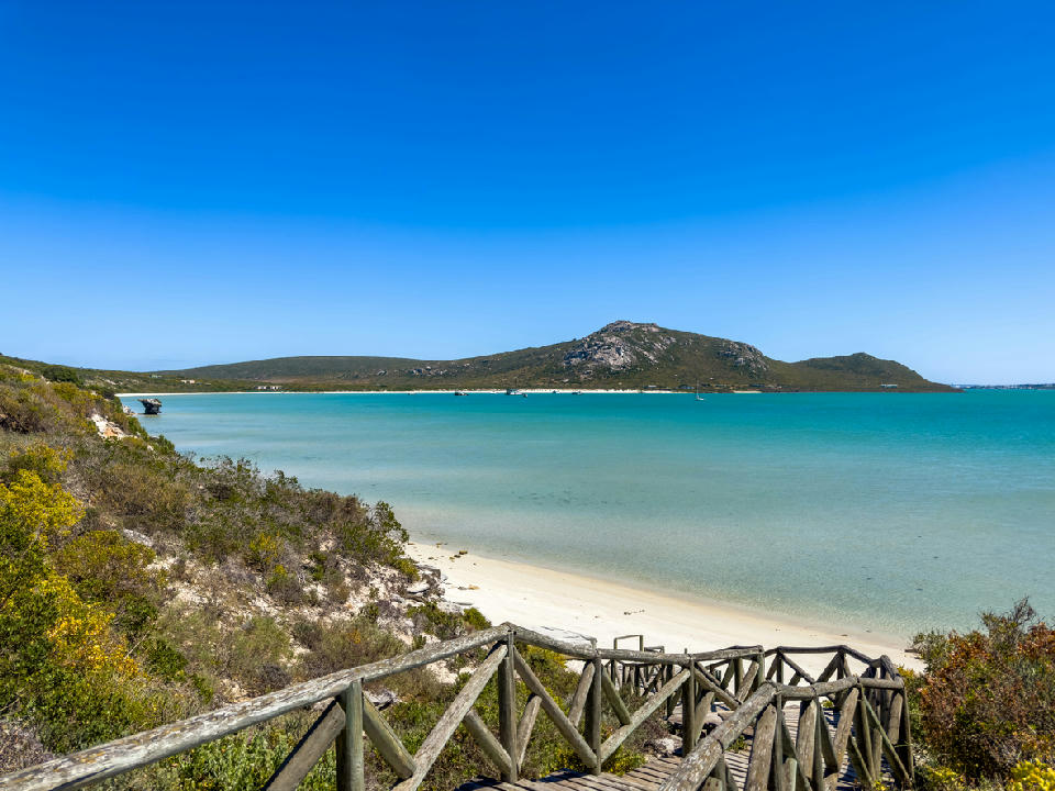 Wooden boardwalk descending to a white sand beach on Langebaan Lagoon with turquoise water and coastal hills in the background.