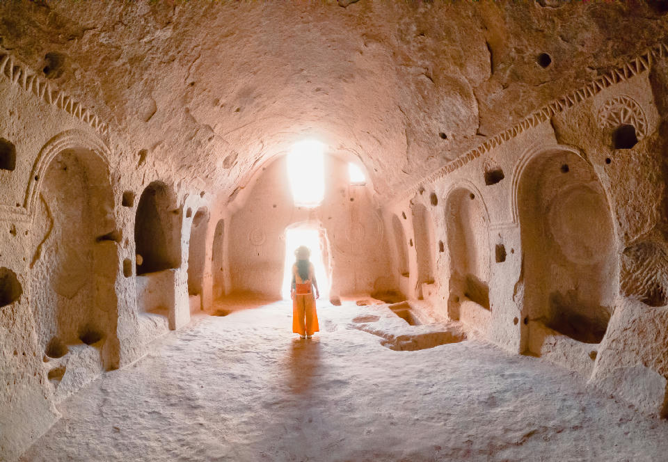 Interior of a rock-cut church at Zelve Open Air Museum in Cappadocia, Turkey, with carved arches, niches, and a visitor silhouetted in the doorway.