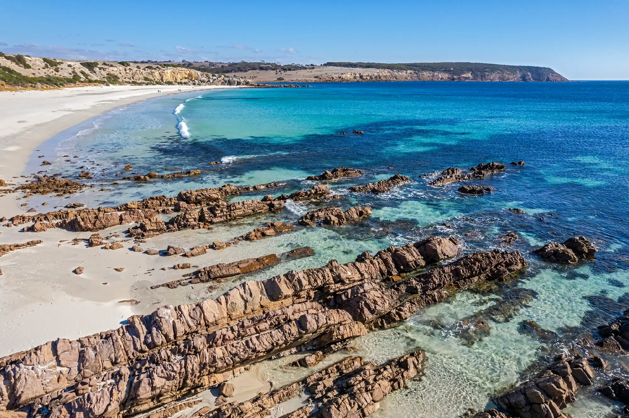 Elevated view of Stokes Bay on Kangaroo Island, showing layered coastal rocks, turquoise shallows, a sweeping white-sand beach and distant cliffs.