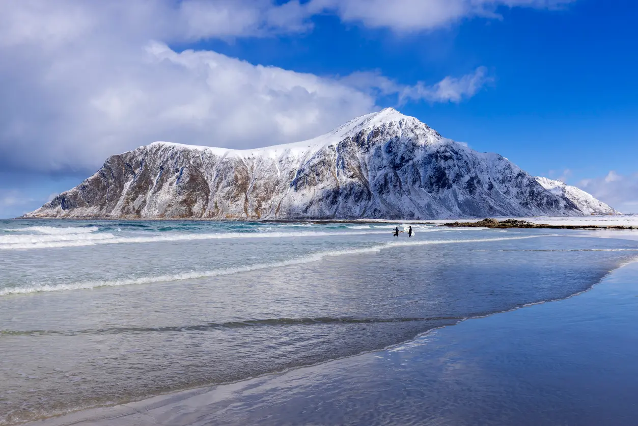 Snow-covered mountains rising above Unstad Beach on Vestvågøya in the Lofoten Islands, with winter waves along the sandy shoreline in Nordland, Norway.