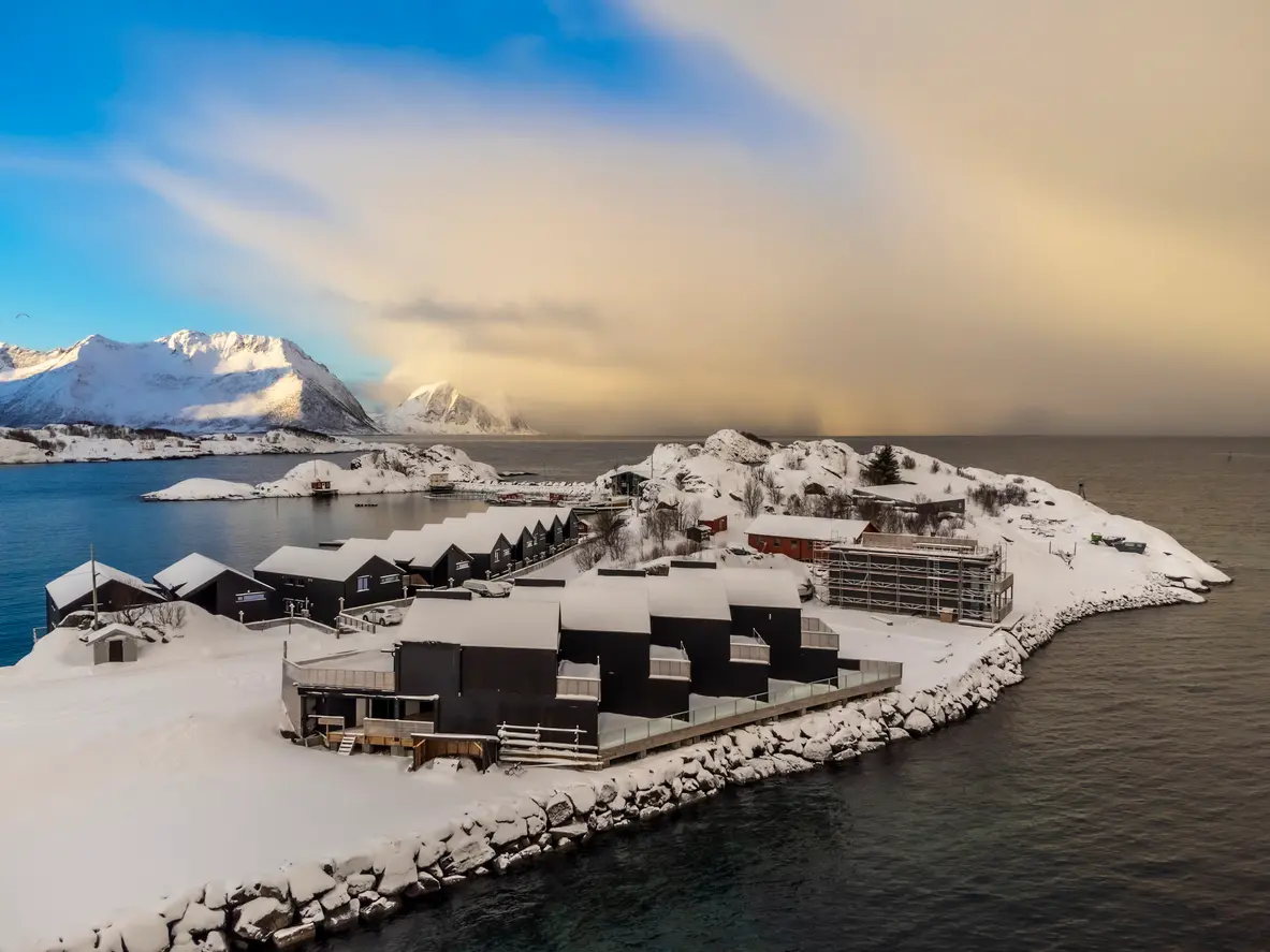 Elevated view of black rorbu cabins and harbour at Hamn i Senja surrounded by snow-covered rocky shoreline and open sea