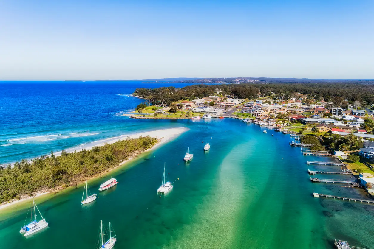 Aerial view over turquoise Currambene Creek with sailing boats and a sandy spit, opening into Jervis Bay beside the waterfront town of Huskisson.