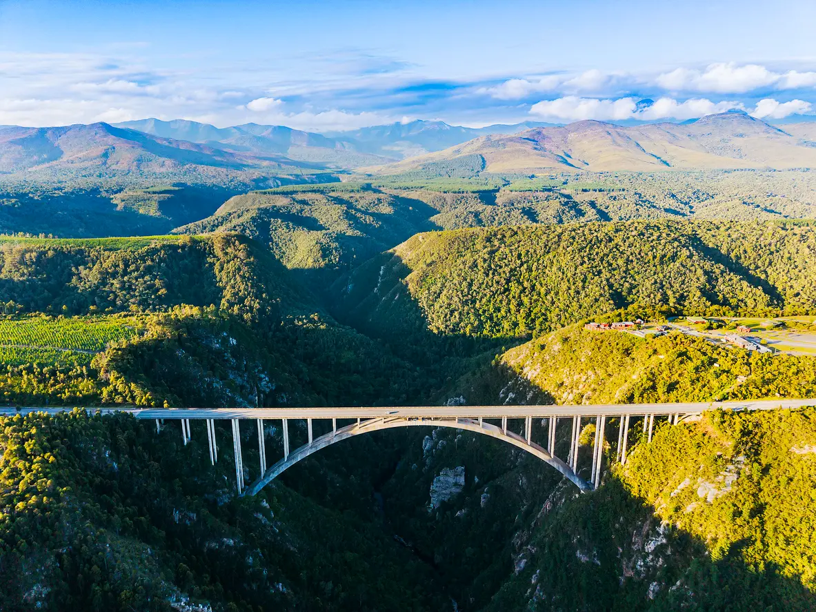 Aerial view of Bloukrans Bridge crossing a deep forested gorge in the Tsitsikamma region of South Africa, with mountain landscapes stretching across the Garden Route hinterland.