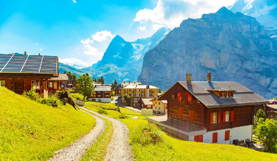 Mürren village with chalets and a winding path across green slopes, overlooking steep cliffs and high alpine peaks in the Swiss Alps.
