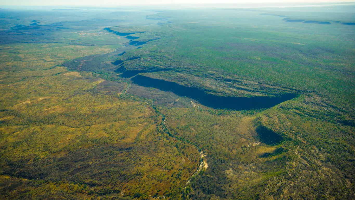 Aerial view of a winding river beneath a long sandstone escarpment surrounded by vast, remote woodland on the Mitchell Plateau.