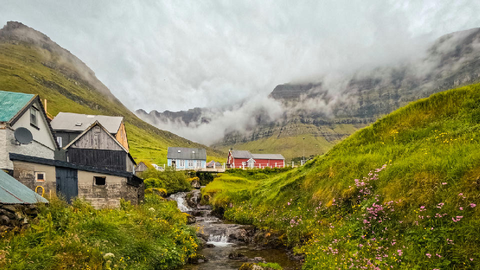 Small houses in Kunoy village beside a stream flowing through a narrow green valley under low cloud and basalt mountains on Kunoy, Faroe Islands.