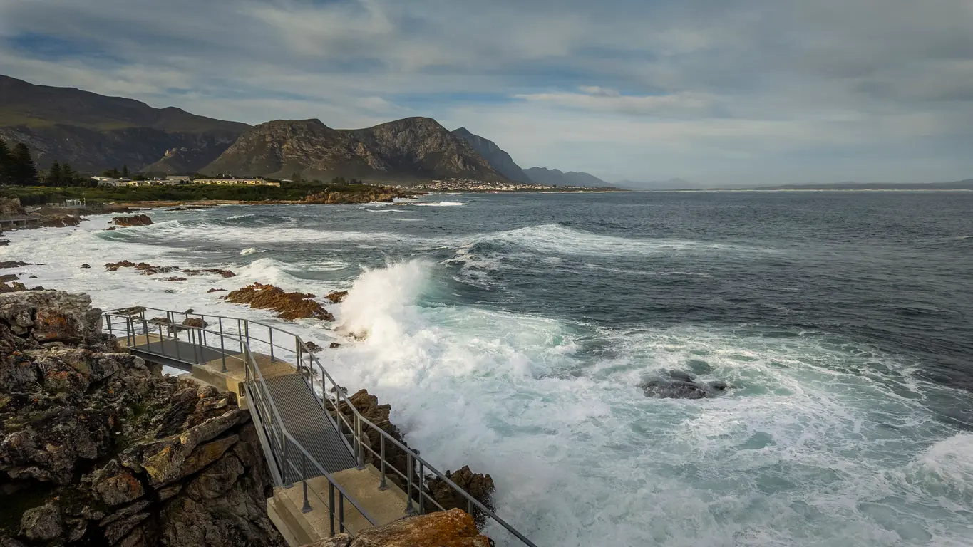 Scenic viewing platform along the Hermanus Cliff Path with crashing ocean waves, rugged coastal rocks, Walker Bay, and mountain scenery in the Overberg region of South Africa.