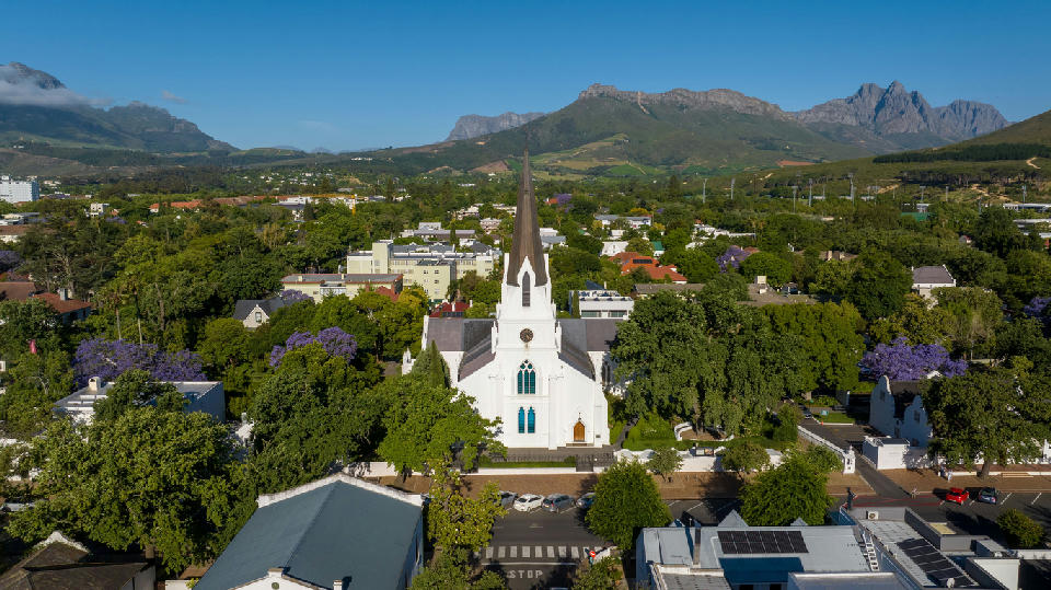 Drone view of Stellenbosch Town Centre featuring the historic white Dutch Reformed Church, tree-lined streets, and Cape Winelands mountains in South Africa.