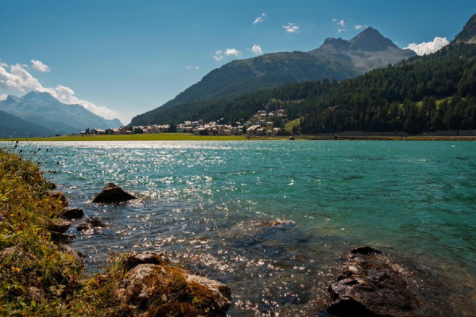 Lake Silvaplana with clear turquoise water, shoreline rocks, and alpine village backed by forested mountains in the Engadin, Switzerland