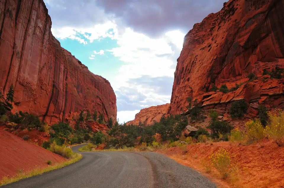 Burr Trail Road curves between towering red sandstone cliffs and desert vegetation beneath dramatic clouds in Long Canyon.