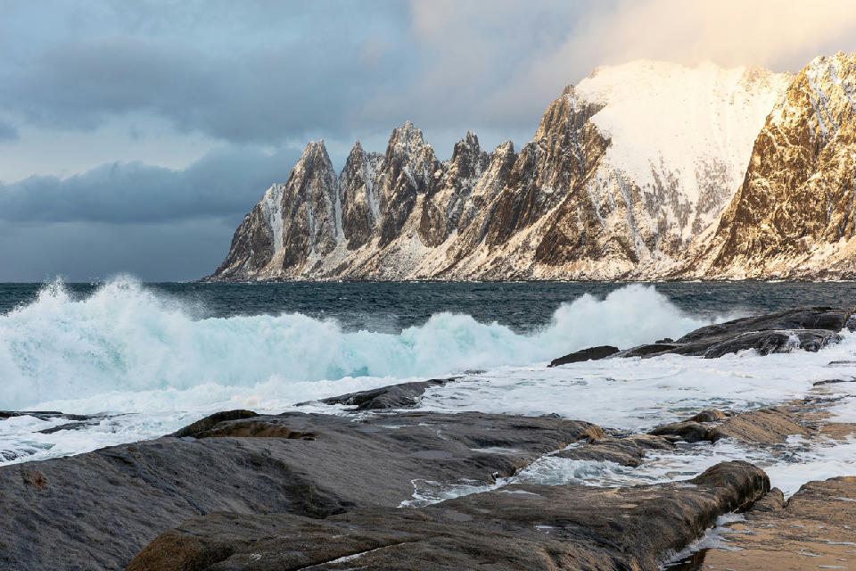 Okshornan Devil’s Teeth peaks Senja Norway from Tungeneset with snow covered jagged mountains and waves crashing over rocky coastline.