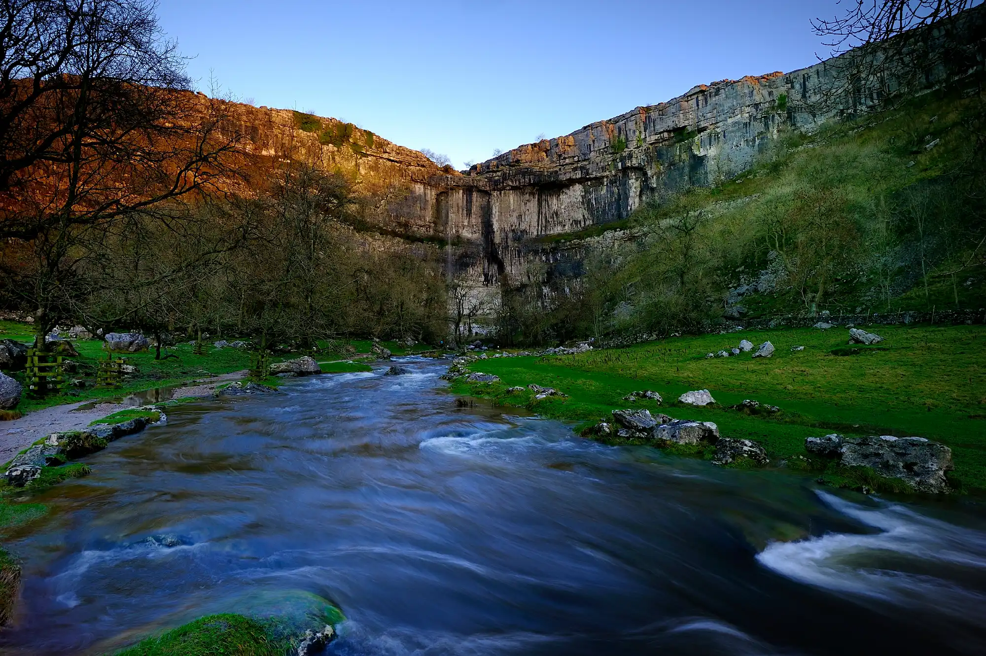 Flowing stream beneath the curved limestone cliffs of Malham Cove, with bare winter trees and warm sunlight on the upper rock face in the Yorkshire Dales.