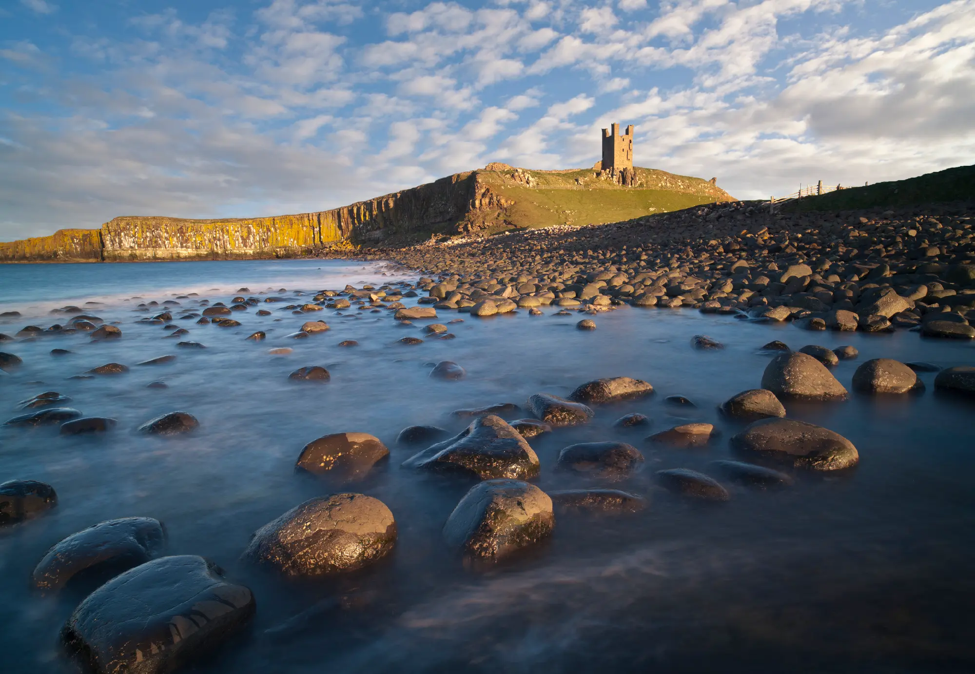 Dunstanburgh Castle tower on a grassy coastal headland above rocky Northumberland cliffs, with smooth long-exposure seawater flowing around foreground boulders in warm afternoon light.