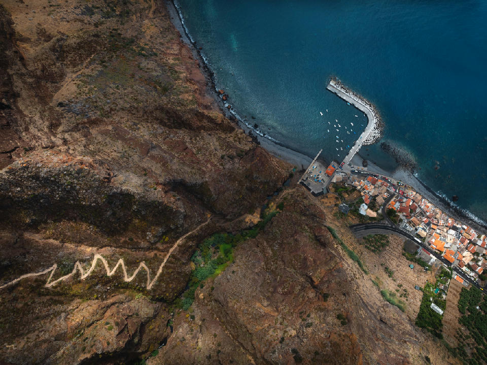 Coastal village of Paul do Mar seen from above, with a steep zigzag path descending the cliffs toward the harbour and Atlantic shoreline.