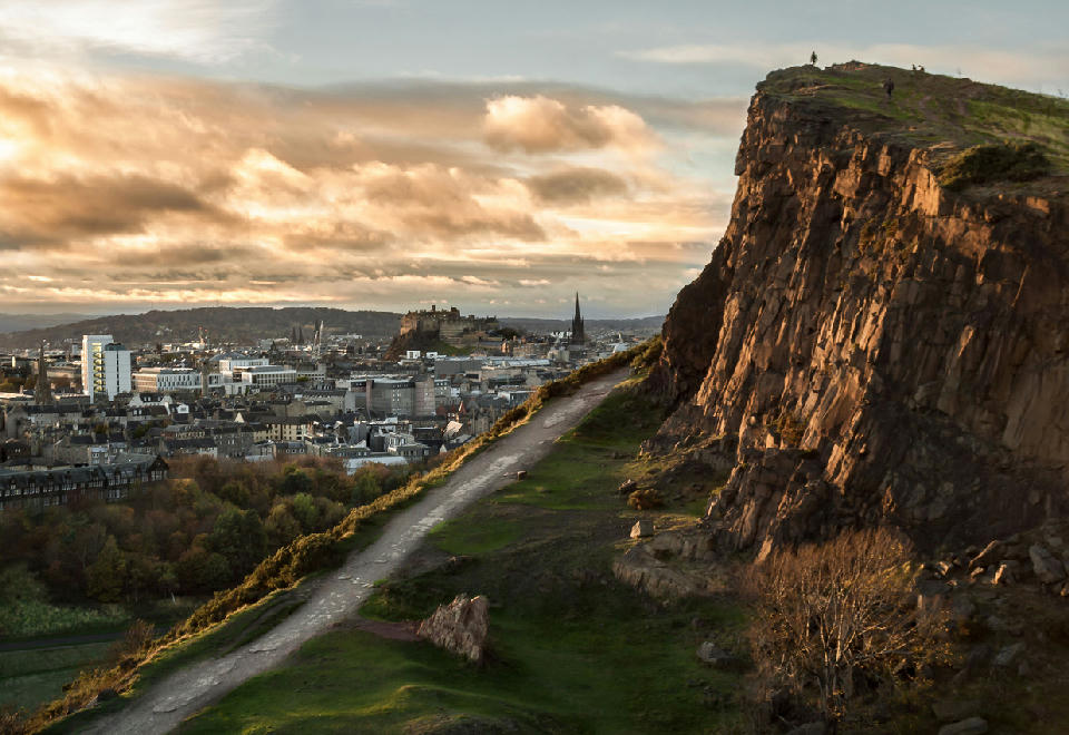 Salisbury Crags cliffs in Holyrood Park with a view toward Edinburgh Castle and the city skyline.