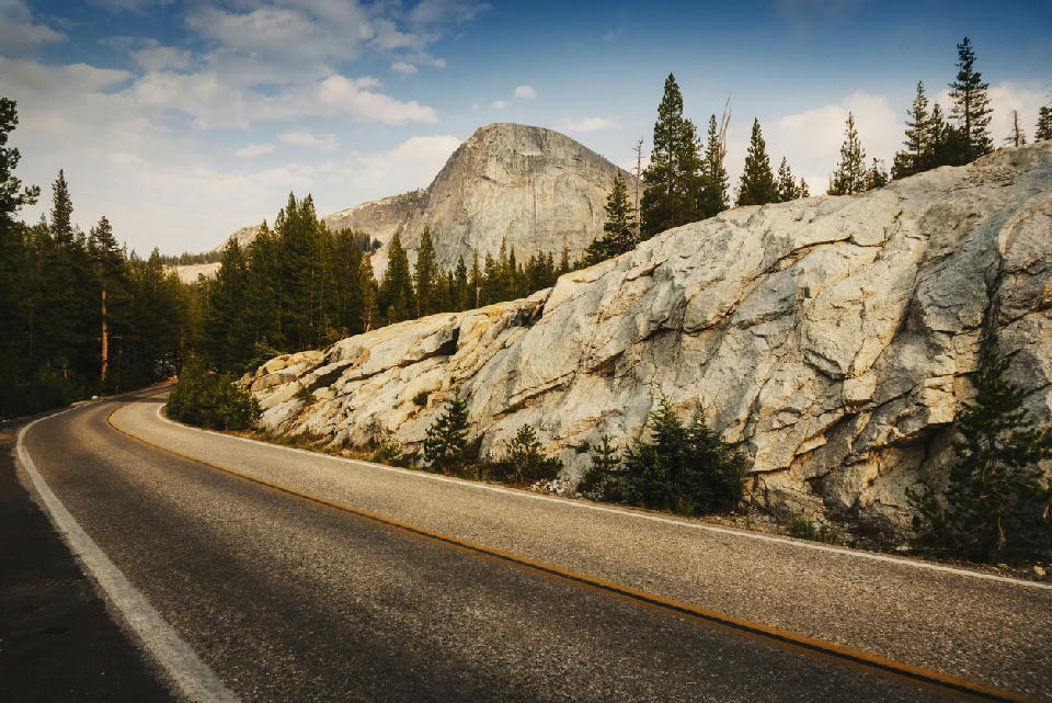 Curving Tioga Pass Road beside exposed granite slabs and conifer forest in Yosemite National Park, California.