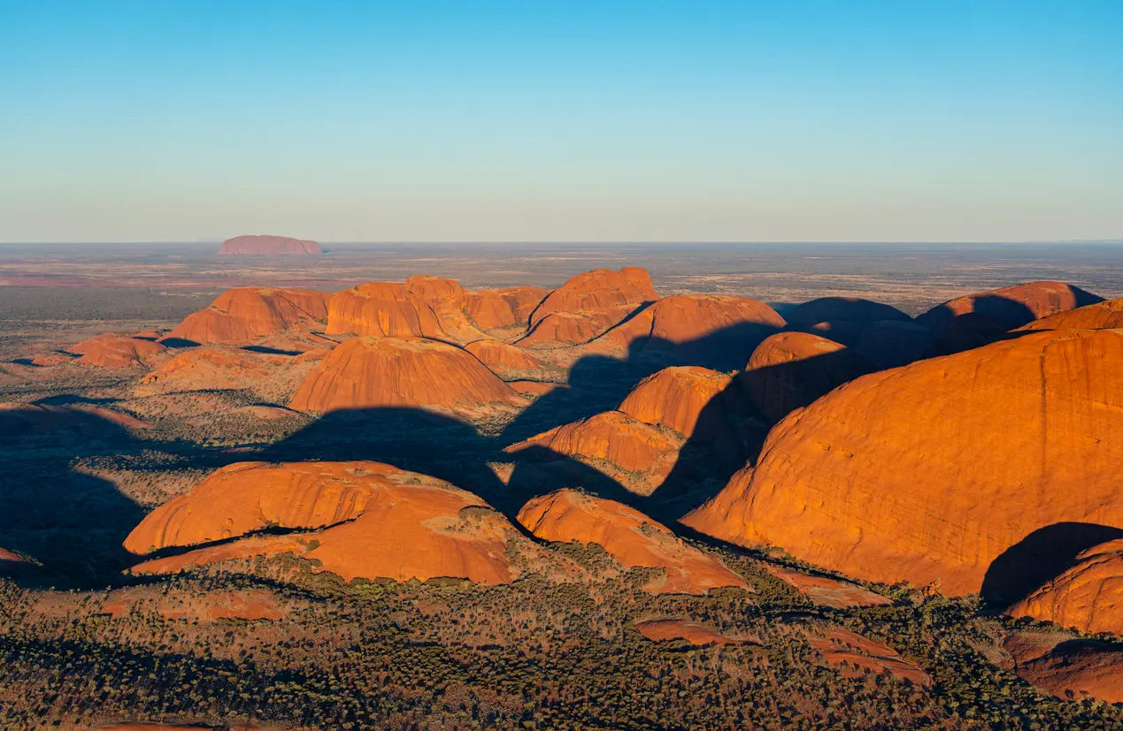 Aerial view of the red sandstone domes of Kata Tjuta in evening light, with Uluru visible on the distant horizon.