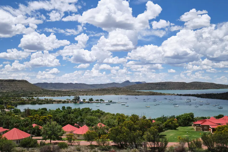 View across Gariep Dam reservoir with sailboats on the water, red-roofed lodges in the foreground and rolling hills beneath towering summer clouds.