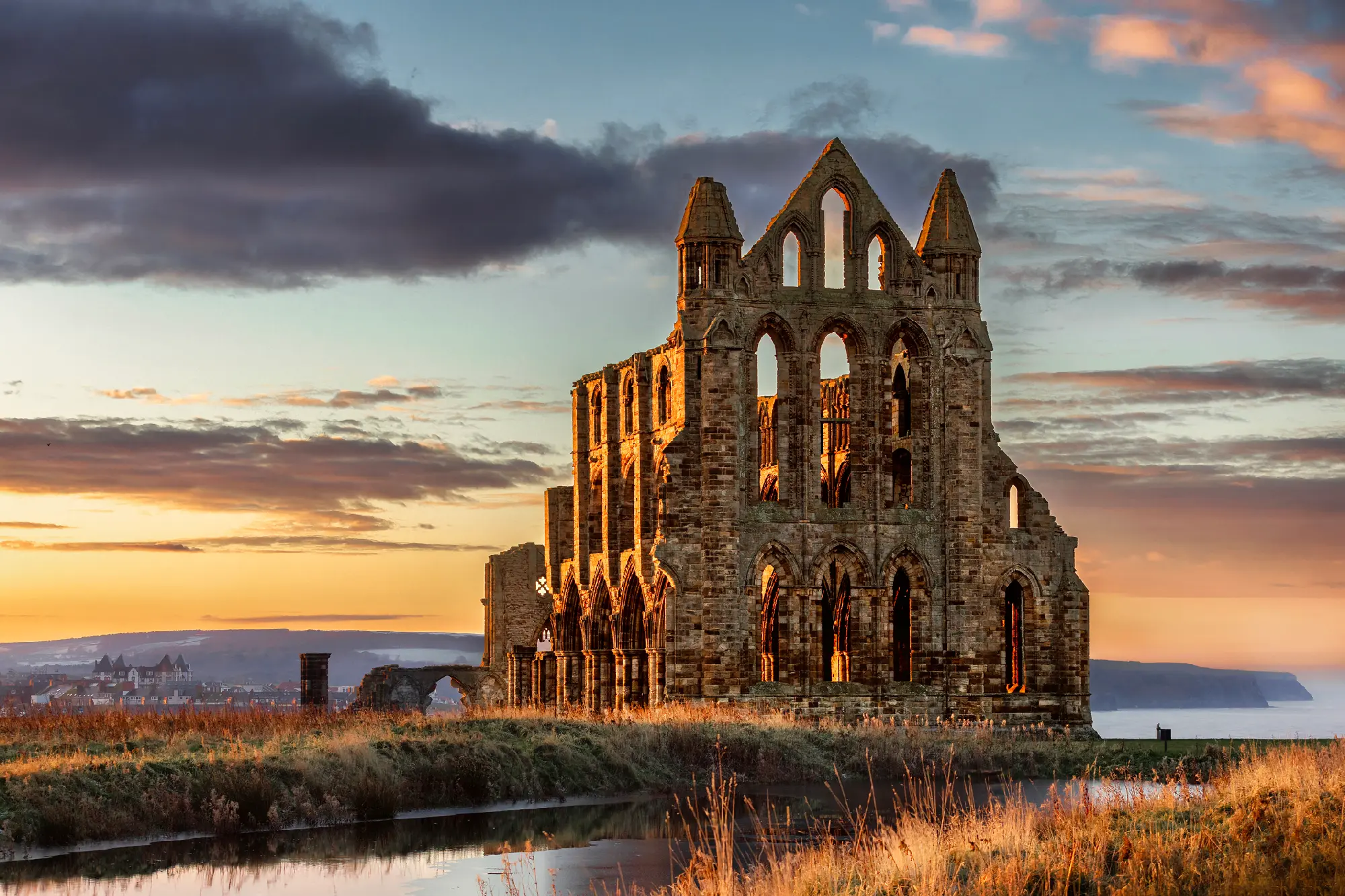 Whitby Abbey ruins illuminated by golden evening sunlight, showing Gothic arches and towers above the North Yorkshire coastline, England.
