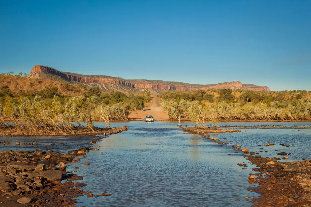 Four-wheel-drive vehicle crossing the shallow Pentecost River with the red cliffs of the Cockburn Range rising in the background.