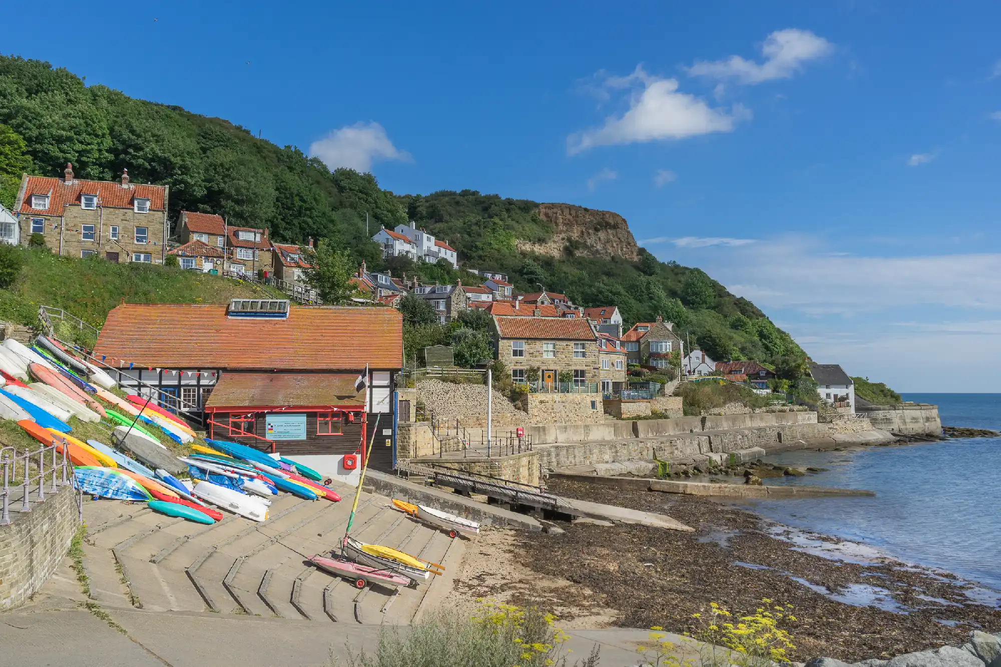 Colourful kayaks beside a waterfront boathouse and slipway at Runswick Bay, with stone cottages, a wooded hillside and the sea beyond.