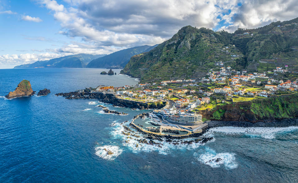 Aerial view of Porto Moniz village on Madeira Island, showing volcanic lava pools, black basalt coastline, and steep green cliffs above the Atlantic Ocean.