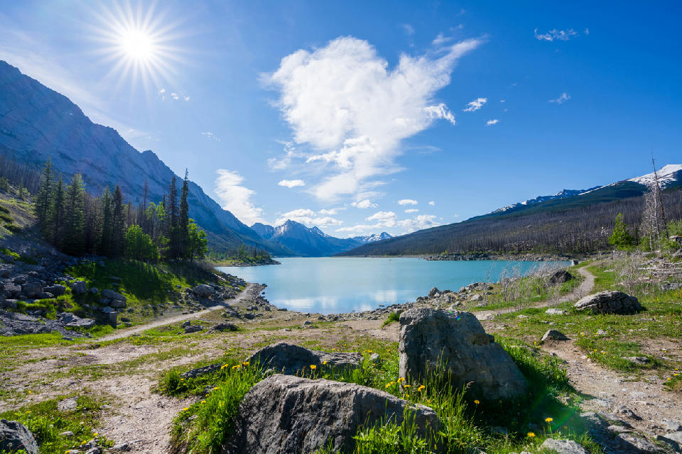 Medicine Lake under bright sun with mountain backdrop, forested shoreline, and rocky foreground path